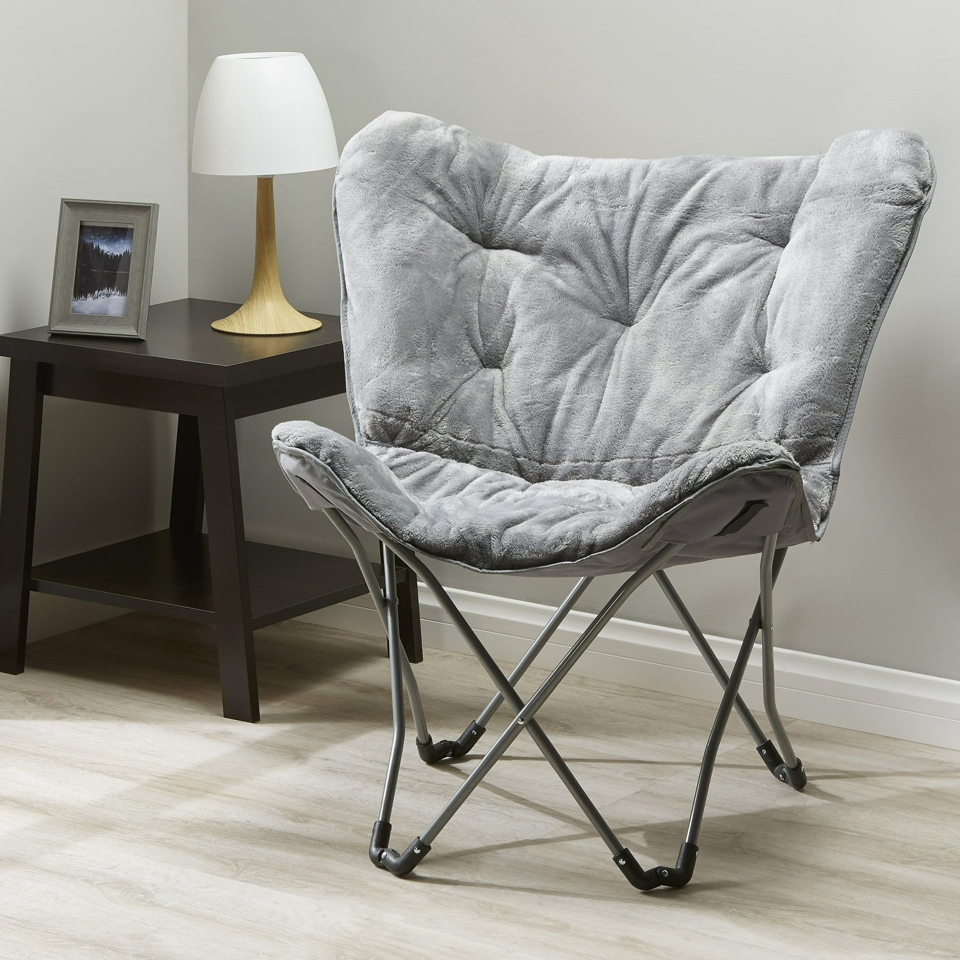 Gray faux-fur butterfly chair with metal legs, shown beside a dark wood side table and white lamp.