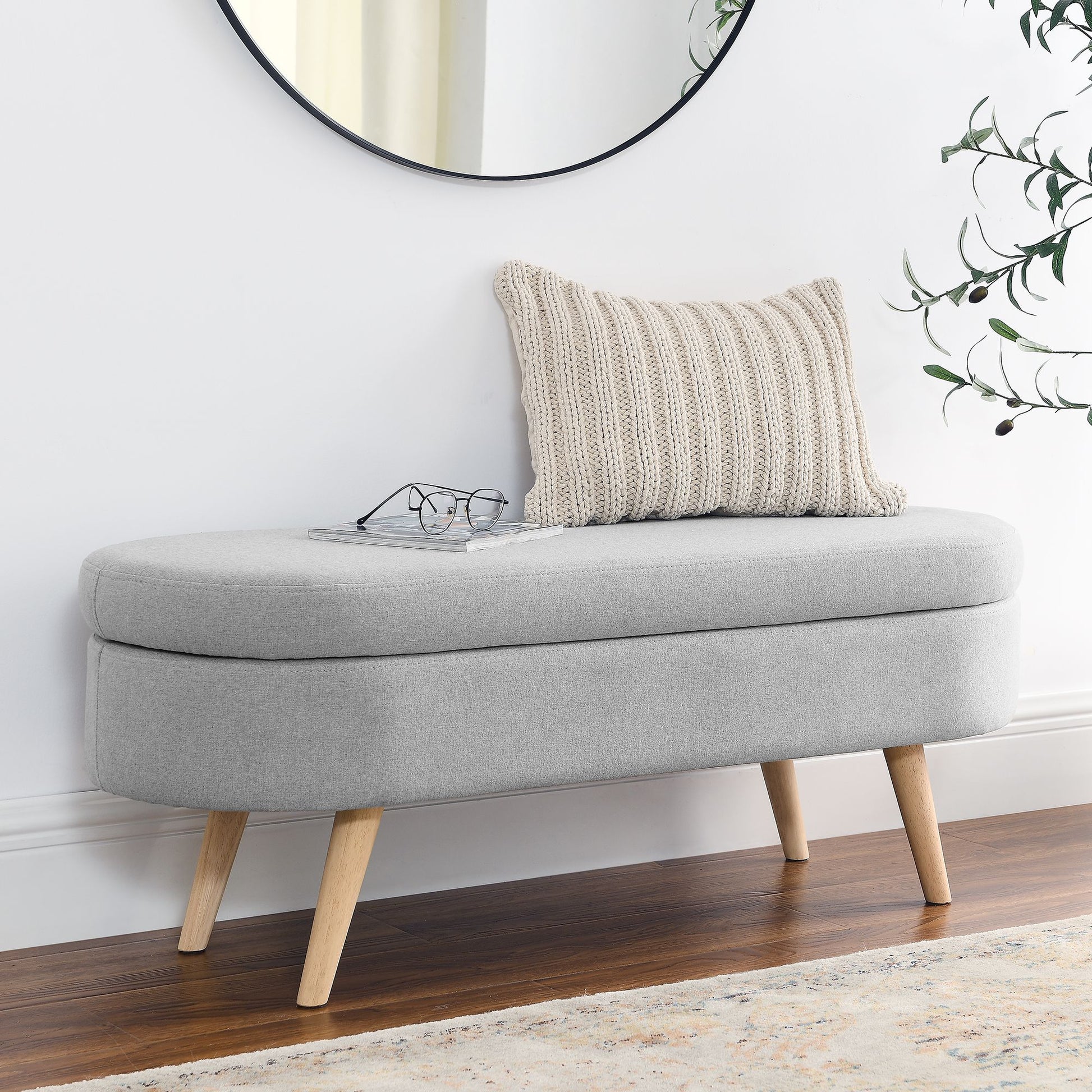 Ottoman oval storage bench with grey fabric upholstery and rubber wood legs.