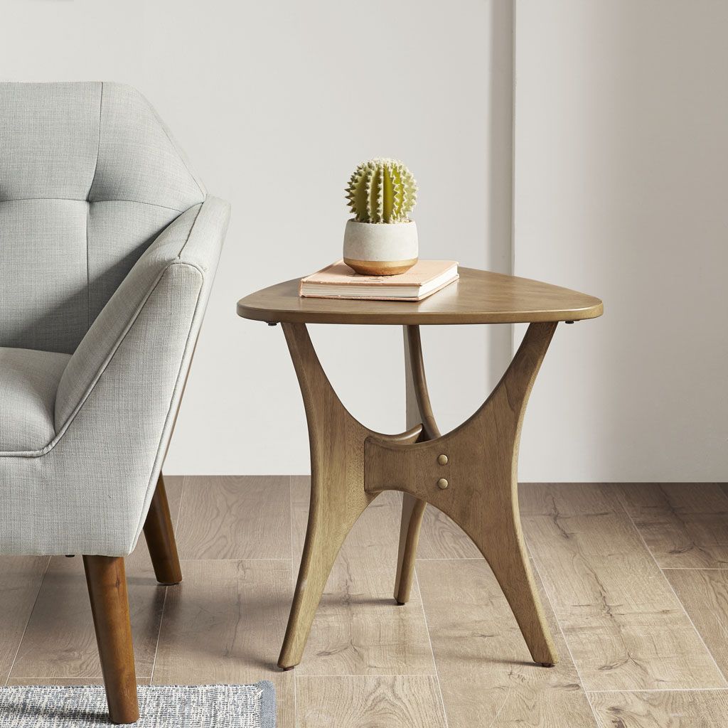 Doba triangle wood side table with natural finish and sculpted curved legs beside a gray upholstered chair.