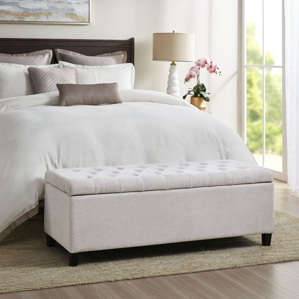 Tufted top soft-close storage bench in light beige fabric with black legs at the foot of a white bed.