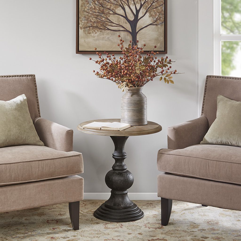 Accent table with round wood top and dark carved pedestal base between two beige armchairs