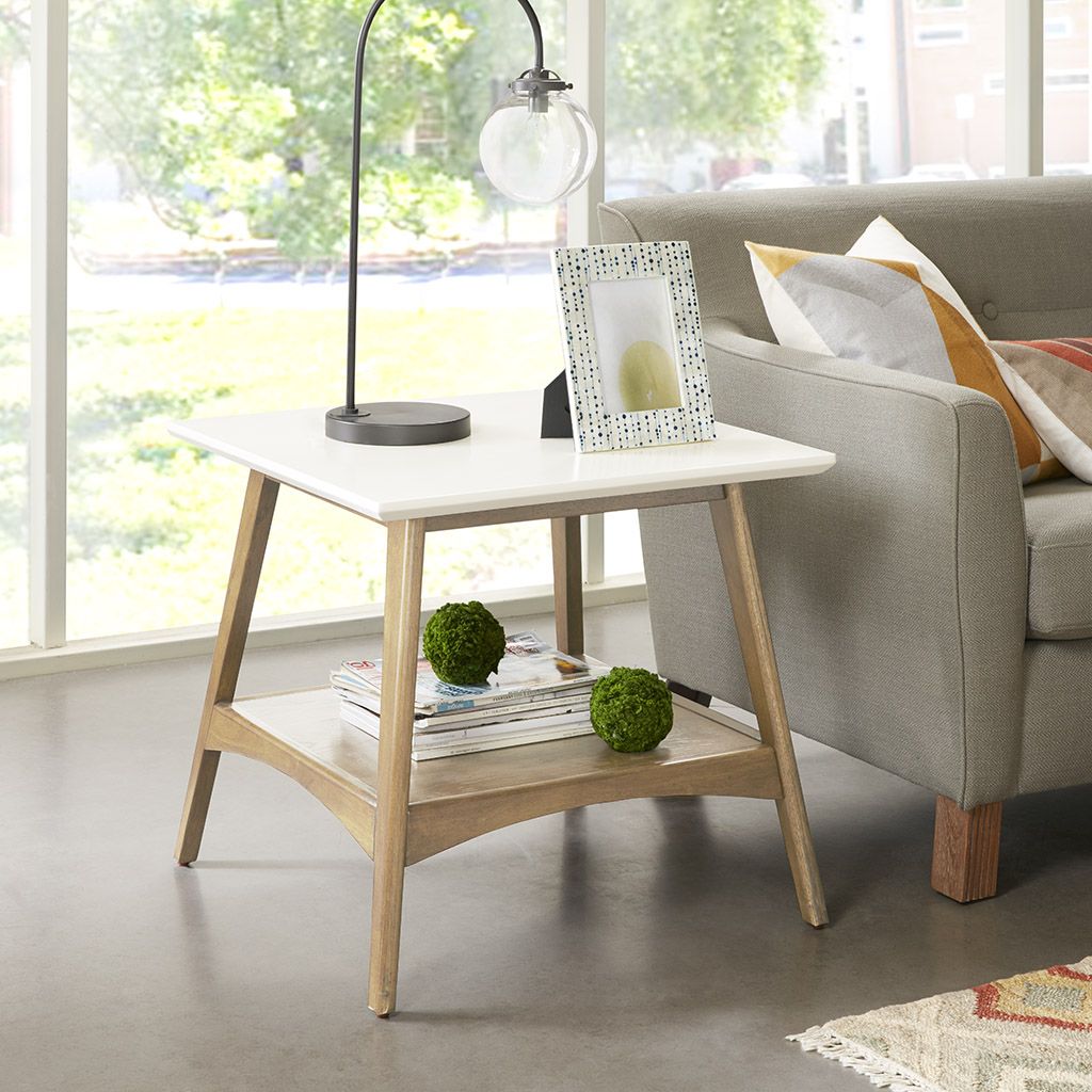 End table with white top and natural wood legs; lamp and photo frame on top, magazines on lower shelf beside gray sofa.
