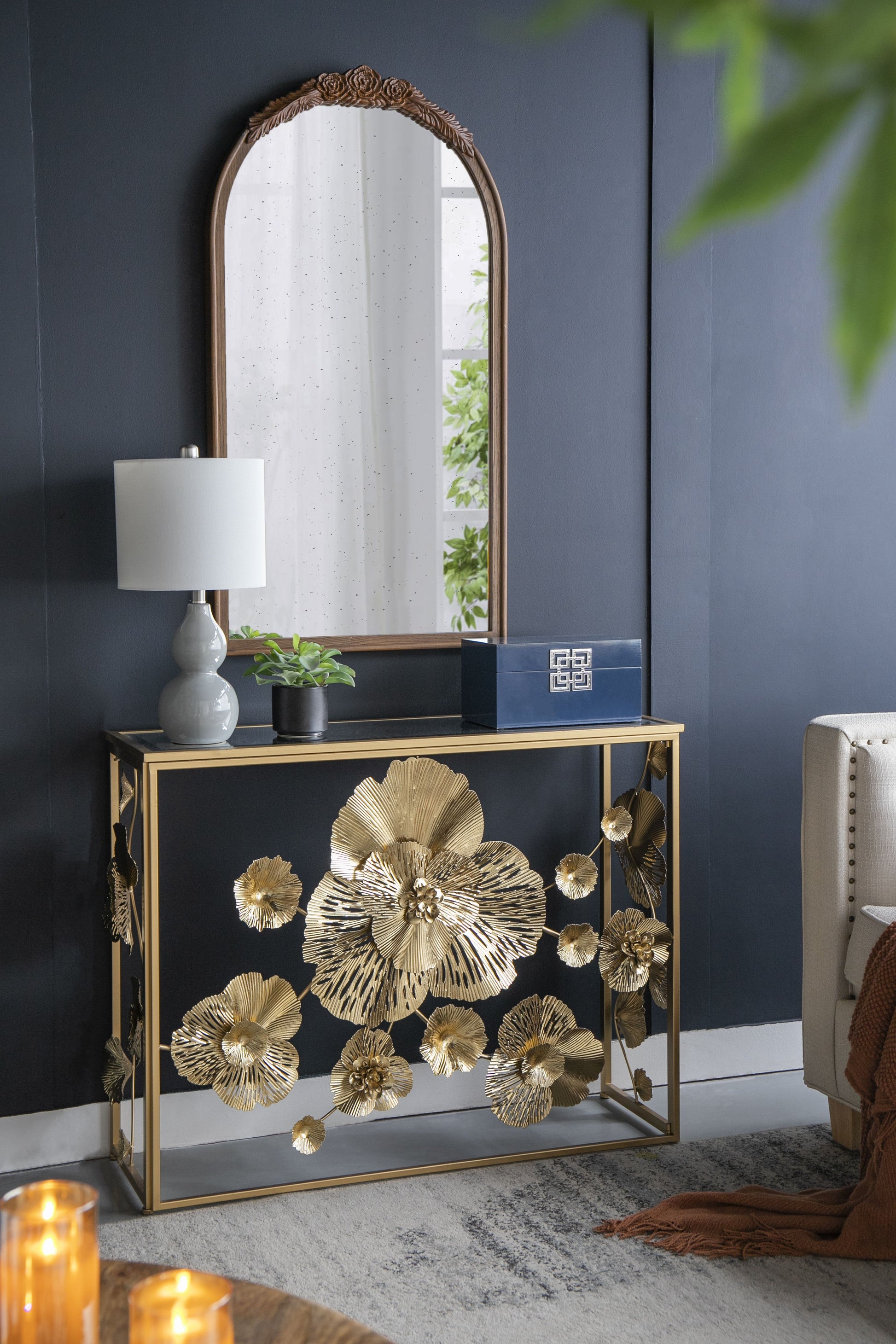 Gold console table with mirrored glass top against a dark blue wall, featuring ornate gold floral base panels