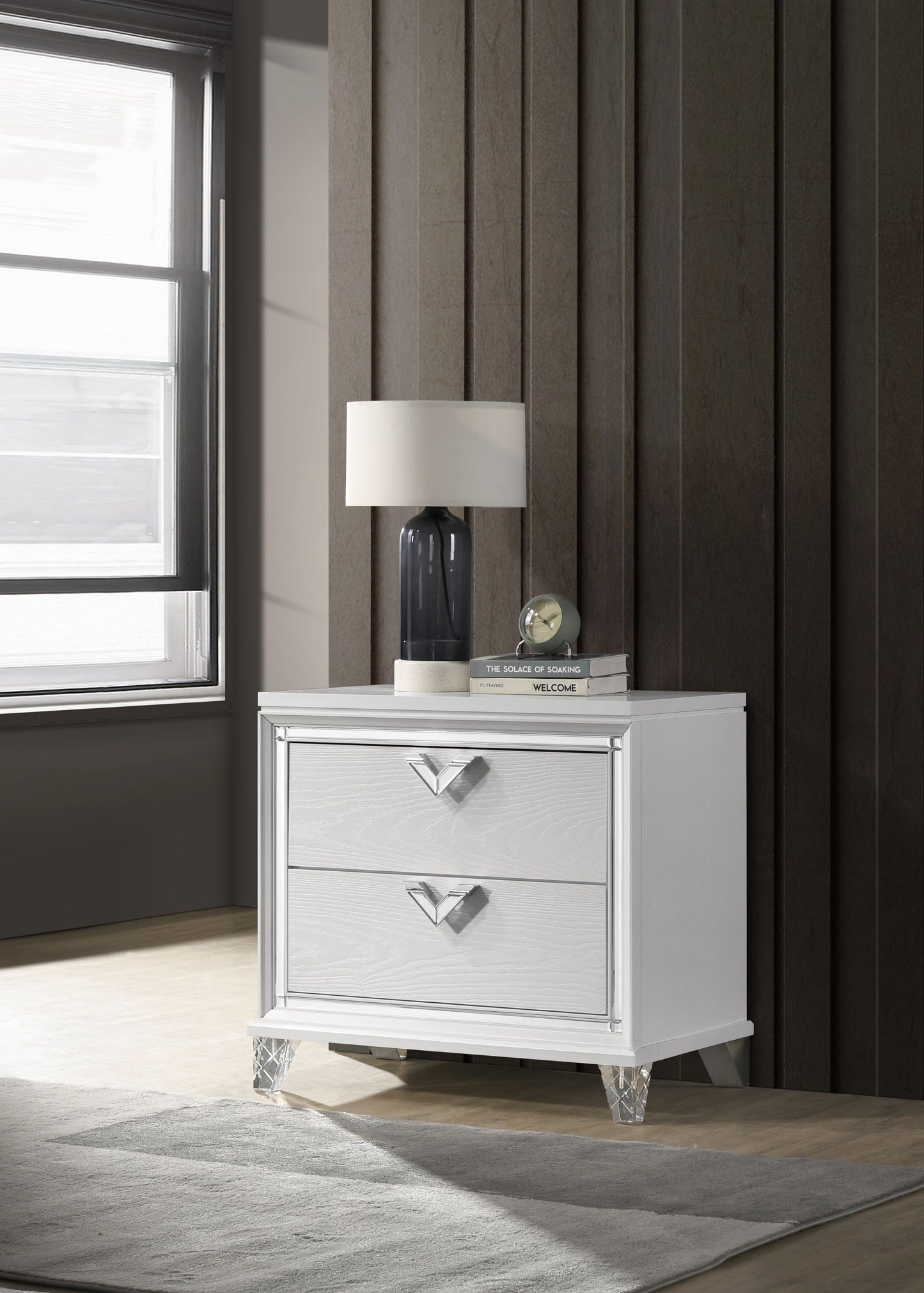Nightstand in white with 2 drawers, V-shaped handles and crystal legs, topped with a lamp and stacked books.