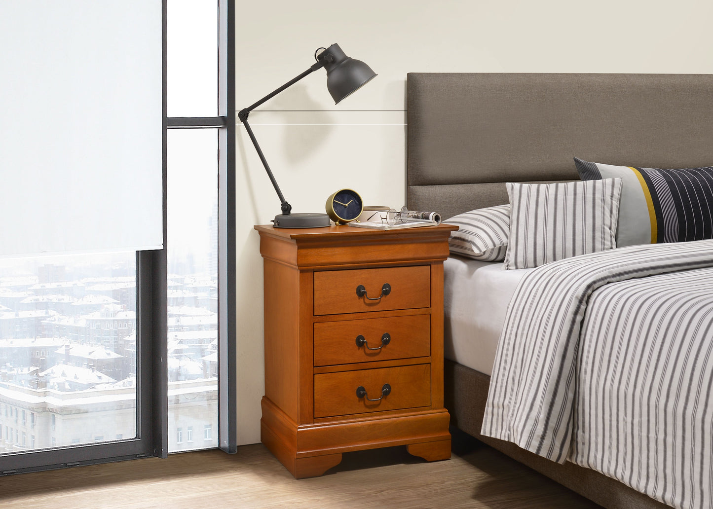Oak-finished nightstand with three drawers beside a bed, topped by a black adjustable lamp.